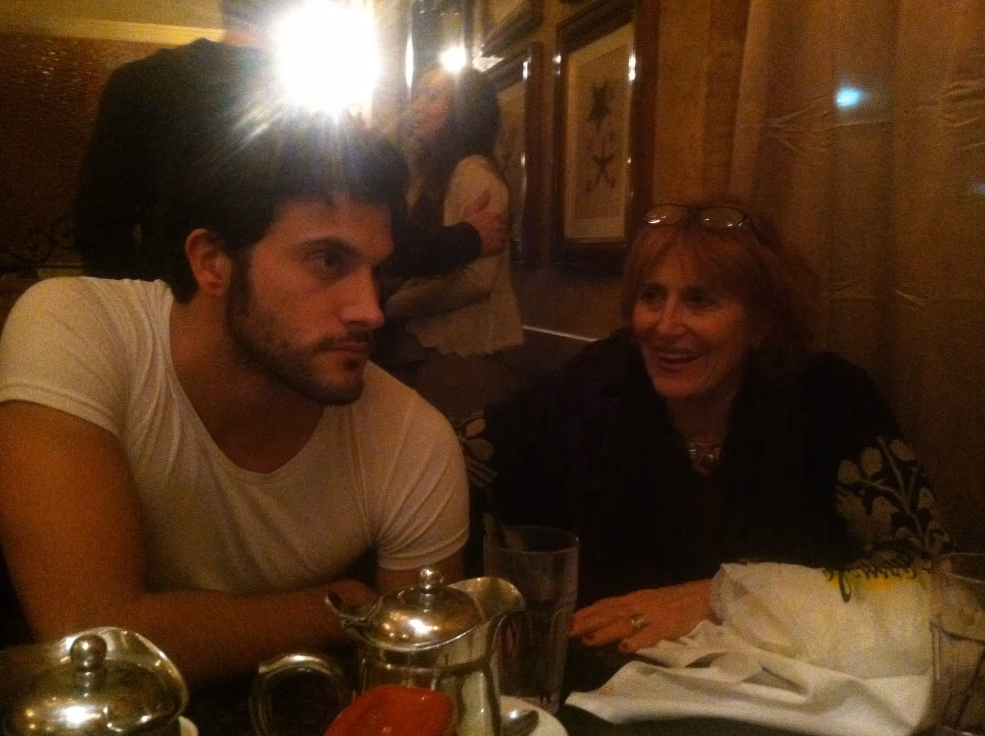 A young man and an older woman seated at a dimly lit dining table with teapots, glasses, and tableware.
