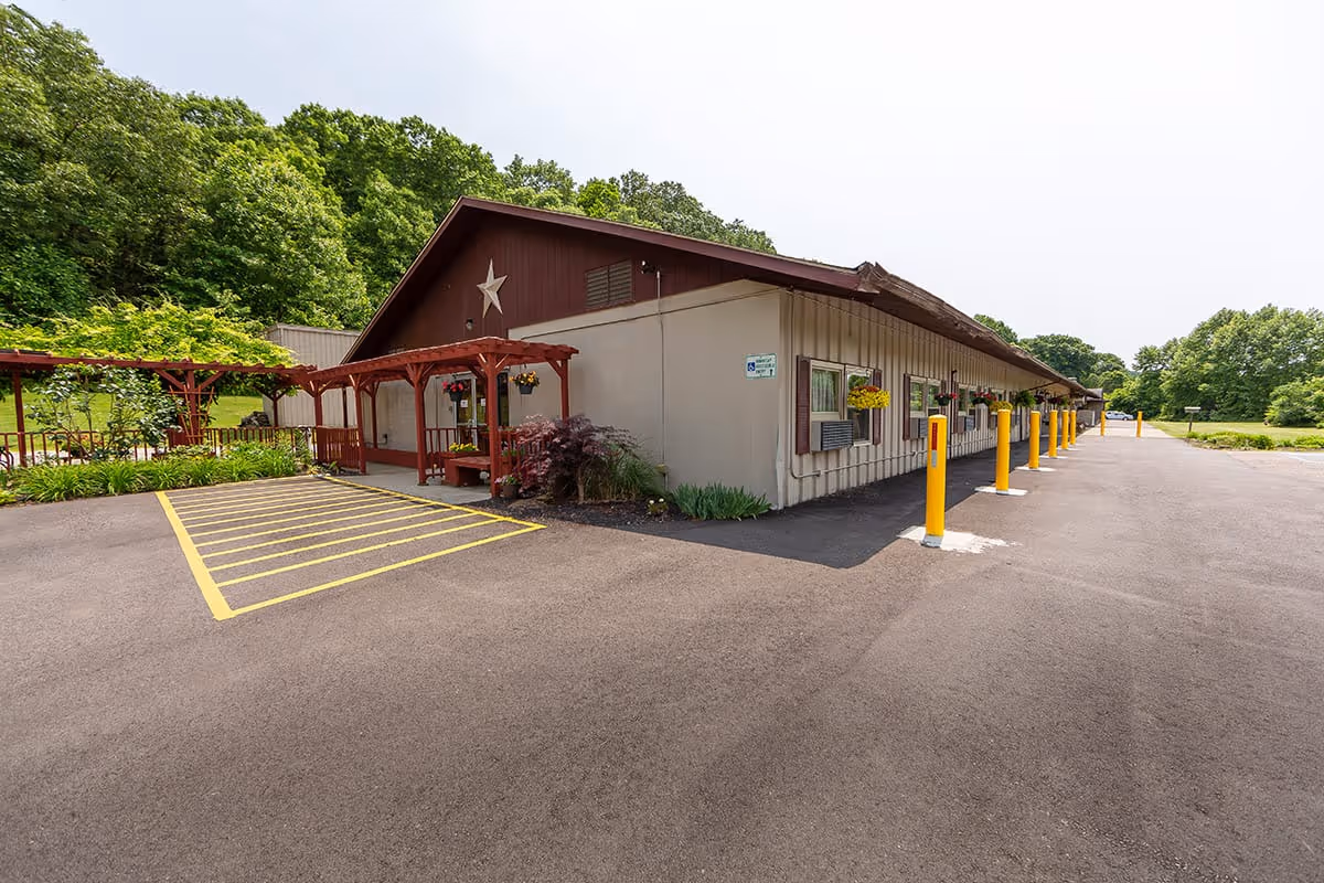 Exterior view of a single-story building with beige siding and a brown roof, featuring a large star decoration on the front. The entrance has a wooden pergola with hanging flower baskets and plants around it. The building is surrounded by a paved parking area with yellow-striped handicap parking spaces and several yellow bollards. Trees and greenery are visible in the background under a cloudy sky.