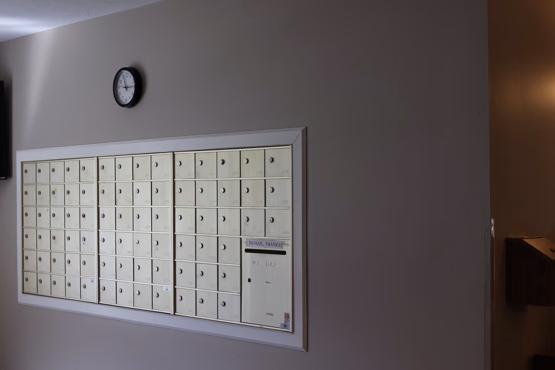 A wall-mounted bank of beige mailboxes with individual locks and numbers, a larger U.S. Mail slot labeled 'TO MAIL, THANKS!' above it, and a round wall clock showing the time as 11:55. The mailboxes are set in a white frame against a light gray wall.