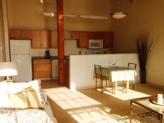 Sunlit interior of an apartment showing a small dining area with a table and two chairs, a kitchen with wooden cabinets, a white refrigerator, microwave, and coffee maker, and part of a living room with a sofa and a lamp. A tall decorative plant is placed near the dining area.