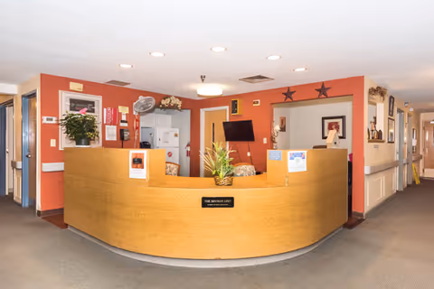 Reception desk area inside Jaffrey Rehabilitation and Nursing Center with a curved wooden counter, a potted plant on the desk, a TV mounted on the wall behind, and hallways leading to other rooms.