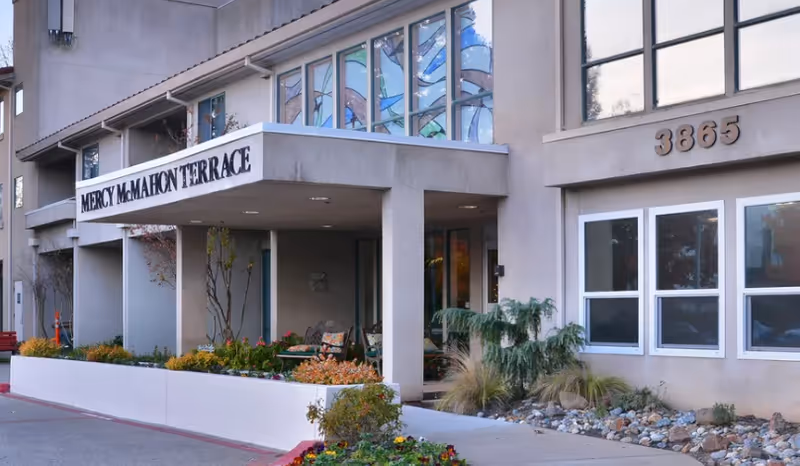 Exterior view of Mercy McMahon Terrace building entrance with a covered porch, decorative stained glass windows above, plants and flowers along the walkway, and the address number 3865 displayed on the wall.