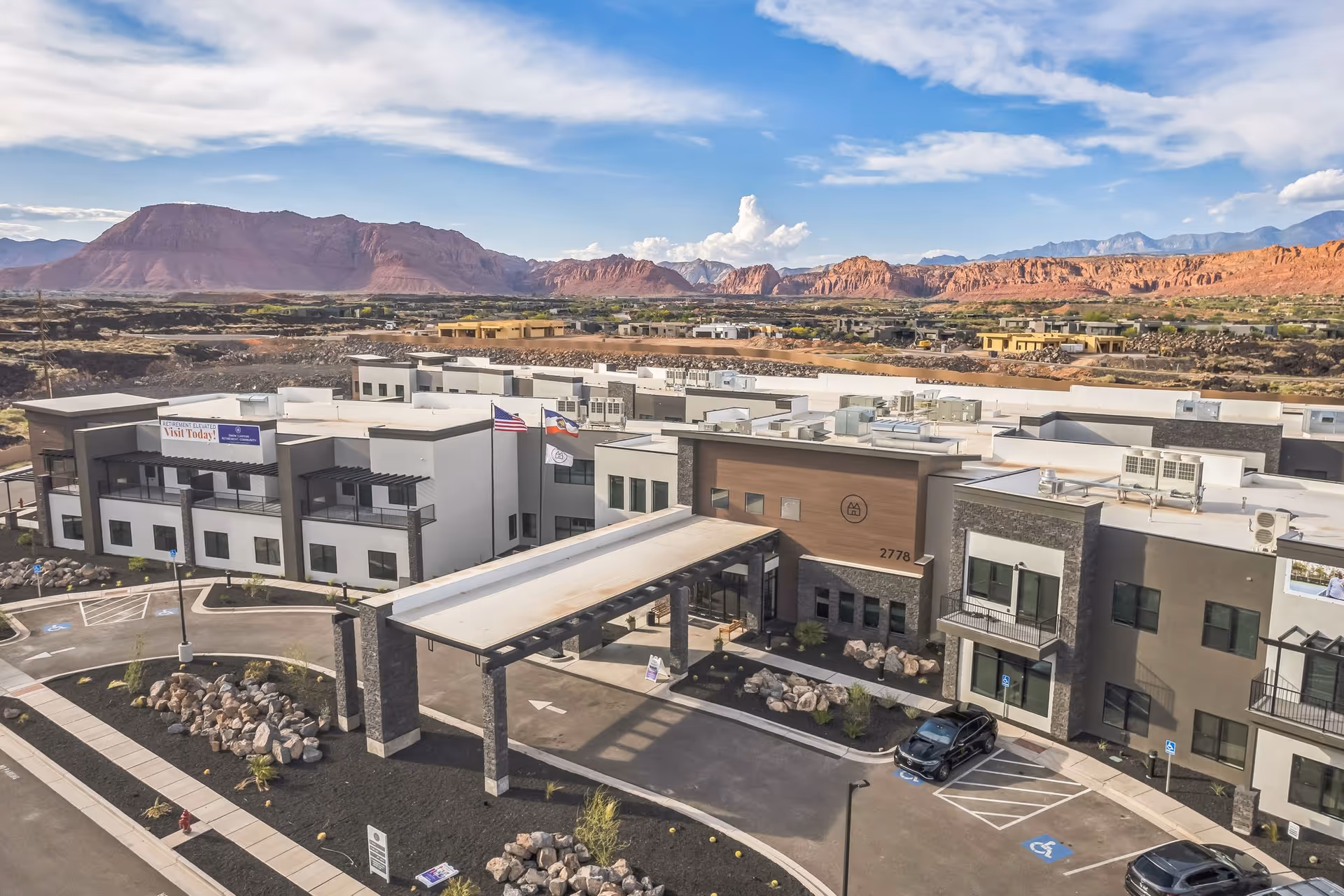 Aerial view of Snow Canyon Retirement Community's main building and covered entrance with parking lot and red rock mountains in the background.