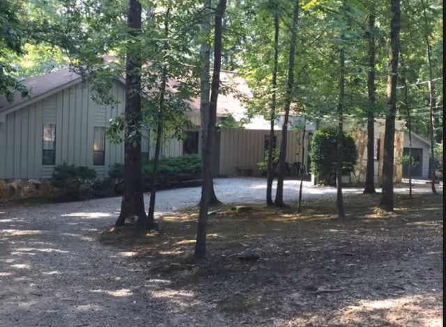 Single-story light-gray building with vertical siding set among trees and a gravel driveway in front.
