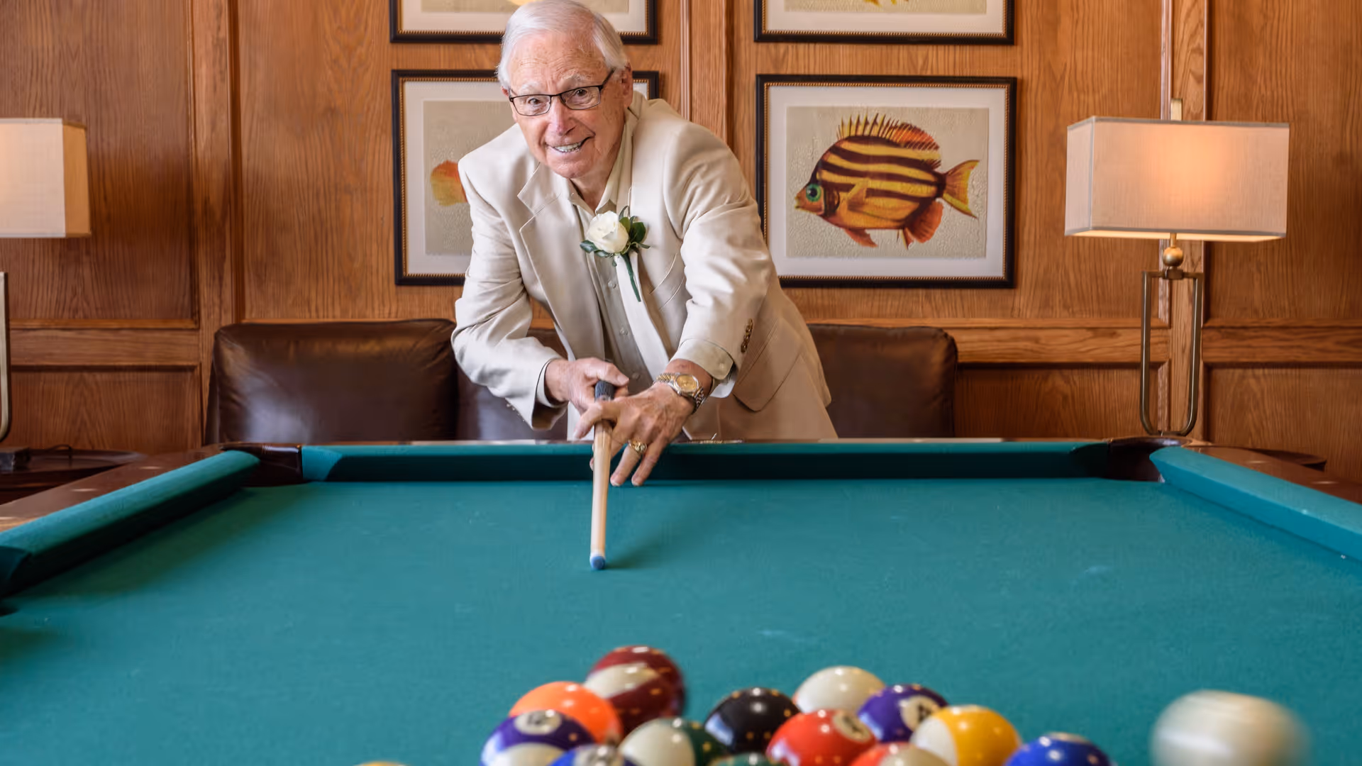 An elderly man wearing glasses and a beige suit jacket is playing pool indoors. He is leaning over a green pool table, aiming with a cue stick. Behind him, there is a brown leather couch, two table lamps with beige shades, and framed pictures of colorful fish on a wooden paneled wall.