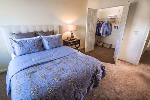 A neatly made bedroom with a blue patterned bedspread, nightstand and lamp, and an open closet with hanging clothes.