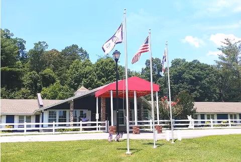 Single-story assisted living building with a red entrance canopy, flagpoles, a white fence and a grassy lawn in front of trees.