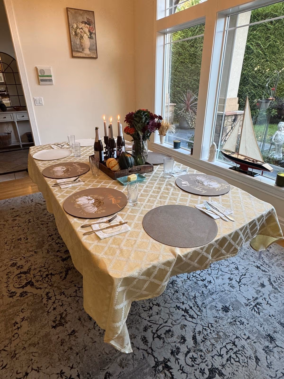 Dining room table set for a meal with placemats, candles, a floral centerpiece and decorative gourds beside large windows overlooking a garden with a model sailboat on the sill.