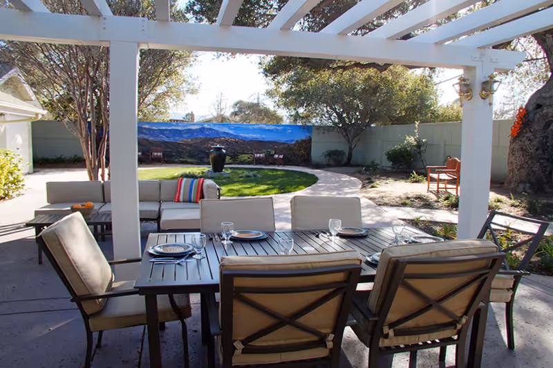 Patio with a pergola-covered dining table and cushioned chairs facing a lawn, mural, and seating area.