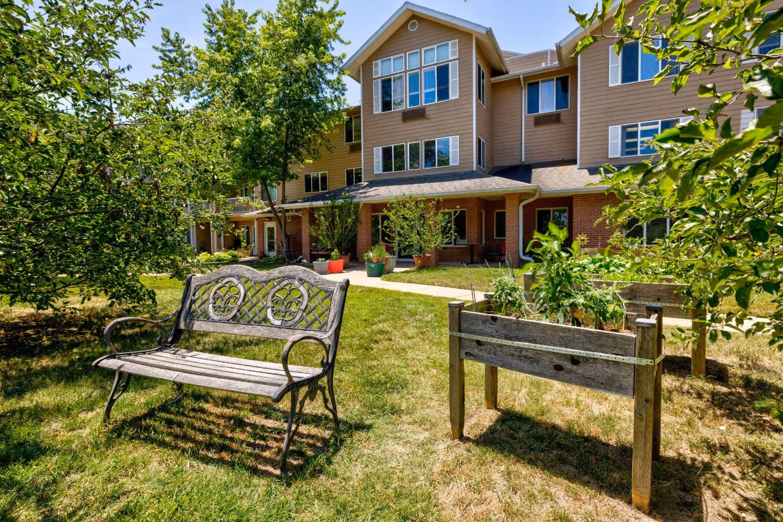 Ornate wooden bench and raised planter boxes on a sunny lawn in front of a multi-story senior living building.