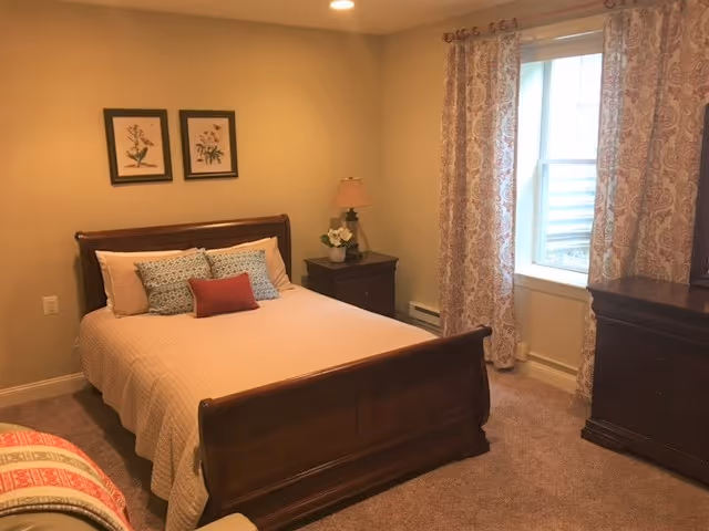 A cozy bedroom with a wooden bed frame, beige bedding, and multiple decorative pillows. There are two framed botanical prints on the wall above the bed. A nightstand with a lamp and a small flower arrangement is next to the bed. A window with patterned curtains lets in natural light, and a wooden dresser is positioned near the window.