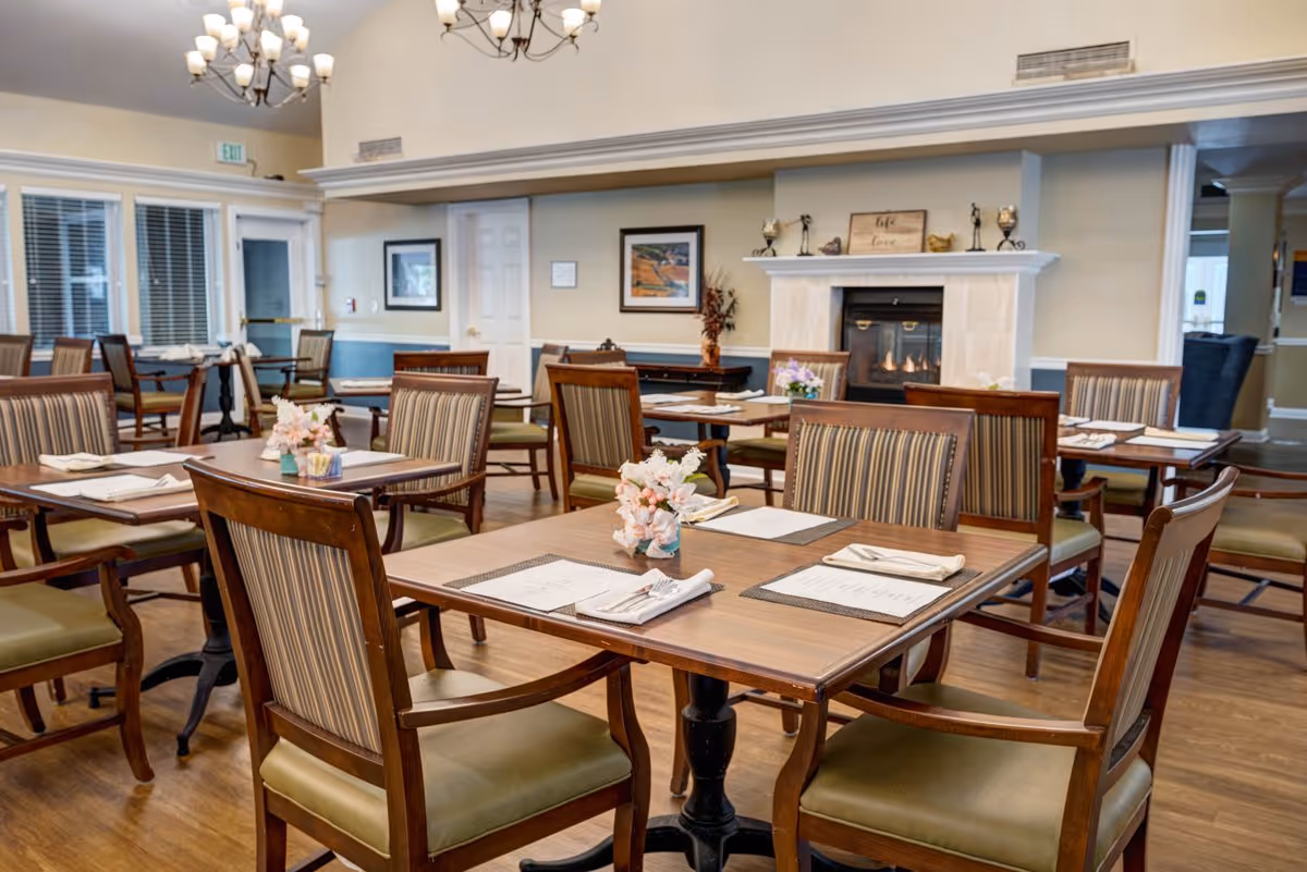 A dining room in a senior living facility with several wooden tables and chairs arranged neatly. Each table has placemats, menus, napkins, and silverware, along with small floral centerpieces. The room features a fireplace with decorative items on the mantel, framed artwork on the walls, and large windows with blinds. The flooring is wooden, and the lighting includes chandeliers hanging from the ceiling.
