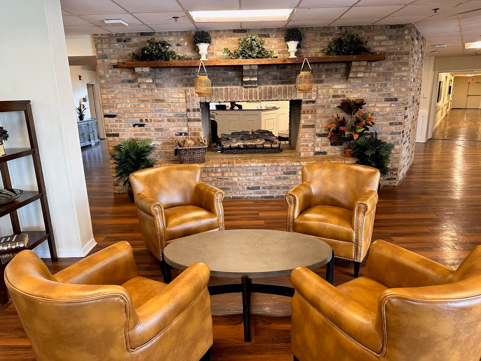 A cozy seating area with four tan leather armchairs arranged around a round coffee table in front of a brick fireplace. The fireplace has decorative plants on the mantel and a basket with logs. The room has wooden flooring and a hallway visible in the background.