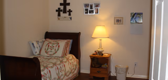 Small bedroom with a single wooden bed covered by a patterned quilt, a nightstand with a lamp, and decorative crosses and photos on the wall.