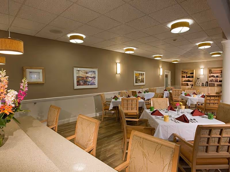 A dining room with several tables covered with white tablecloths, each set with folded burgundy napkins, glassware, and cutlery. The room has wooden chairs with cushioned seats, framed artwork on the beige walls, ceiling lights, and a flower arrangement on a counter in the foreground.