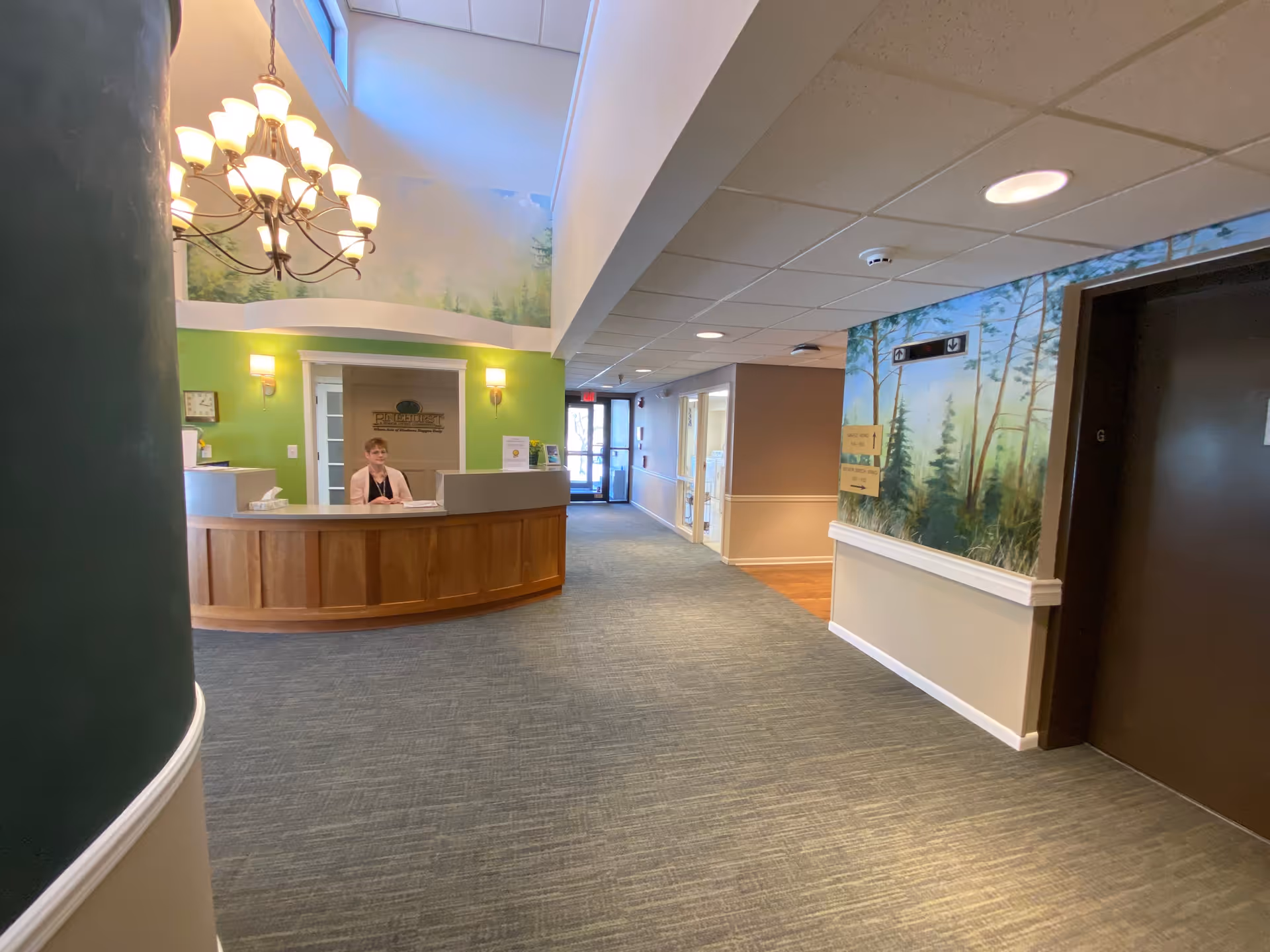 Reception area inside Pinehurst, Episcopal SeniorLife Communities with a curved wooden front desk, a receptionist seated behind it, green walls, a large chandelier, and a mural of trees on the wall near an elevator.