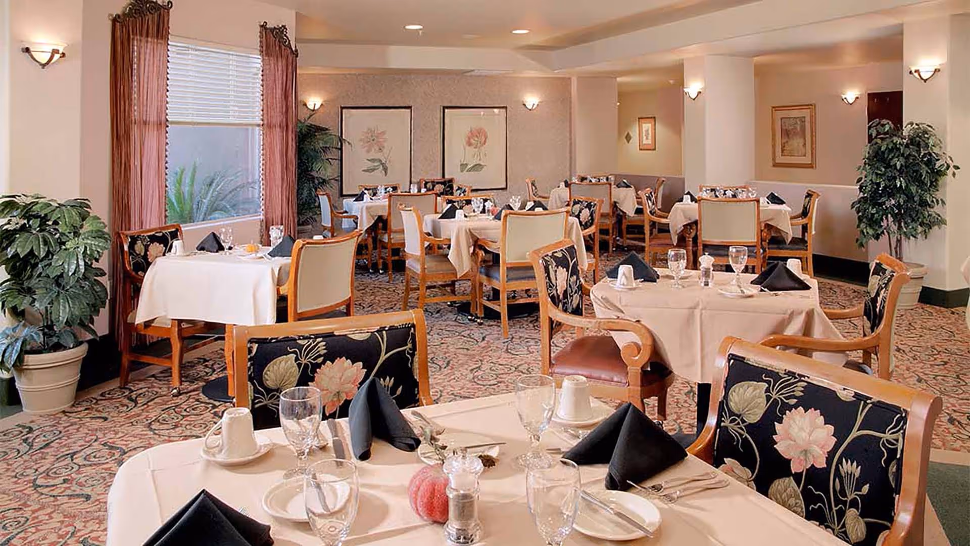 A well-lit dining room with multiple tables covered in white tablecloths, each set with glassware, plates, and black folded napkins. The chairs have floral upholstery, and there are framed floral artworks on the walls. Large windows with sheer curtains allow natural light to enter, and potted plants add greenery to the space.