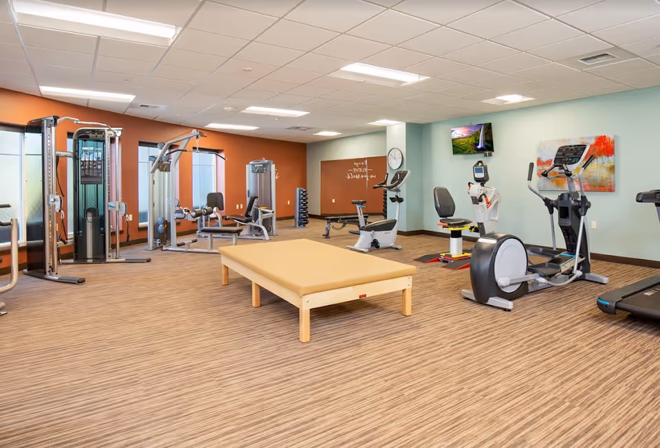 A spacious fitness room with various exercise equipment including a treadmill, stationary bikes, weight machines, and a padded exercise table in the center. The room has light blue and orange walls, a wall clock, a mounted TV, and a colorful abstract painting.