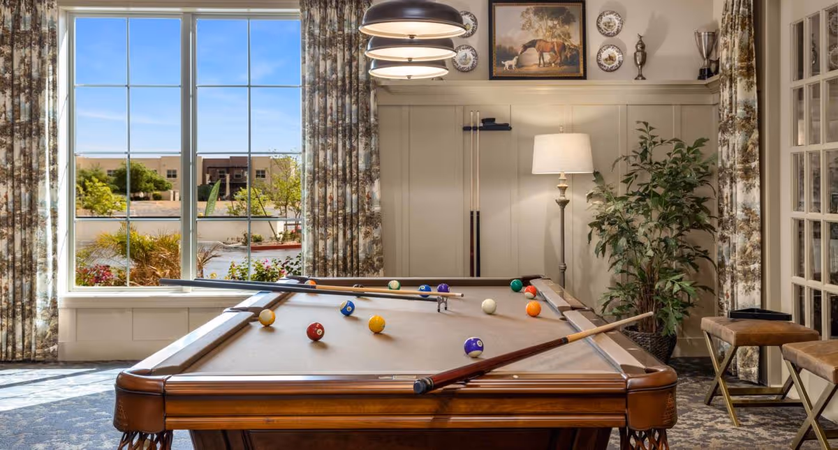 A well-lit interior game room with a pool table, cues, seating, plants and a large window overlooking landscaping.