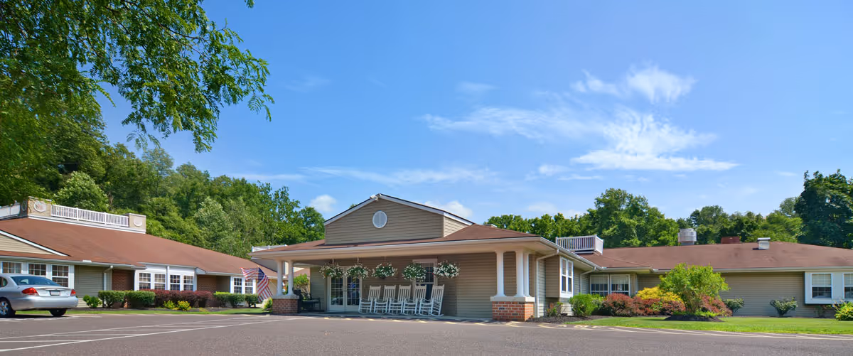 Exterior view of a single-story senior living facility building with a covered entrance featuring several rocking chairs. The building has a brown roof and beige siding, surrounded by green trees and shrubs under a clear blue sky.
