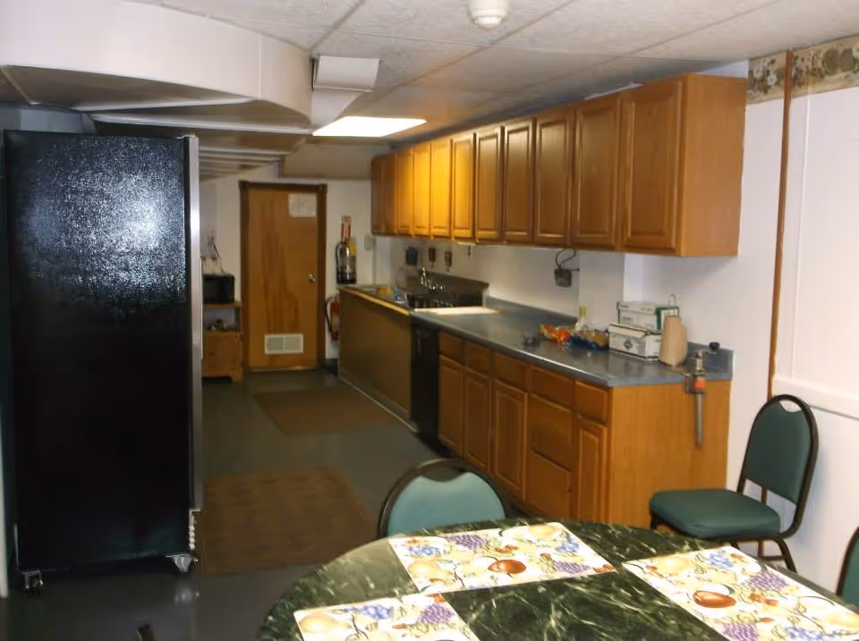 Long narrow communal kitchen with wooden cabinets, a countertop and sink, a black refrigerator, and a table with placemats in the foreground.