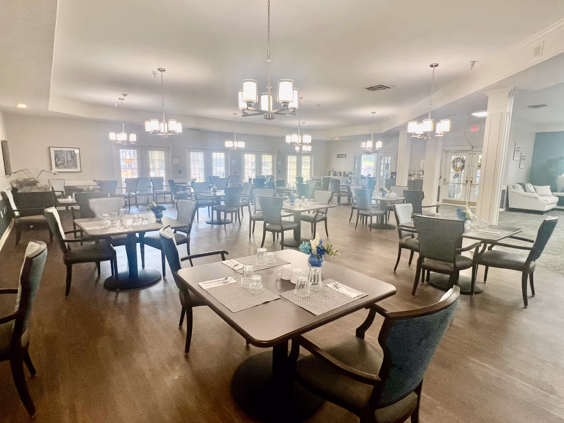 A spacious dining room with multiple tables and chairs arranged neatly. Each table is set with placemats, glasses, and cutlery. The room is well-lit with several modern chandeliers hanging from the ceiling. Large windows and glass doors allow natural light to fill the space. There is a seating area with sofas visible in the background near the entrance.
