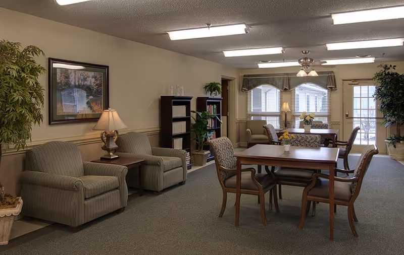 A cozy common area in a senior living facility with several upholstered armchairs, wooden tables with chairs, potted plants, a bookshelf filled with books, a table lamp, framed artwork on the wall, and windows with blinds and valances allowing natural light to enter.