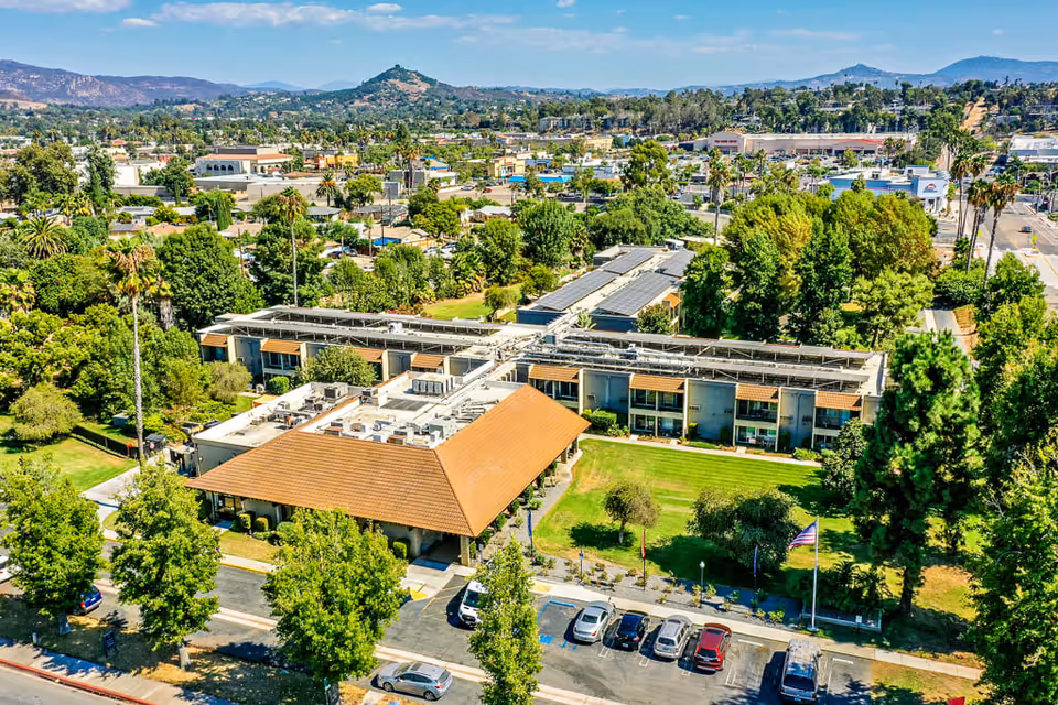 Aerial view of Escondido Senior Living facility showing a large building with a brown tiled roof surrounded by green lawns, trees, and a parking lot with several cars. The background includes a cityscape with hills and mountains under a clear blue sky.