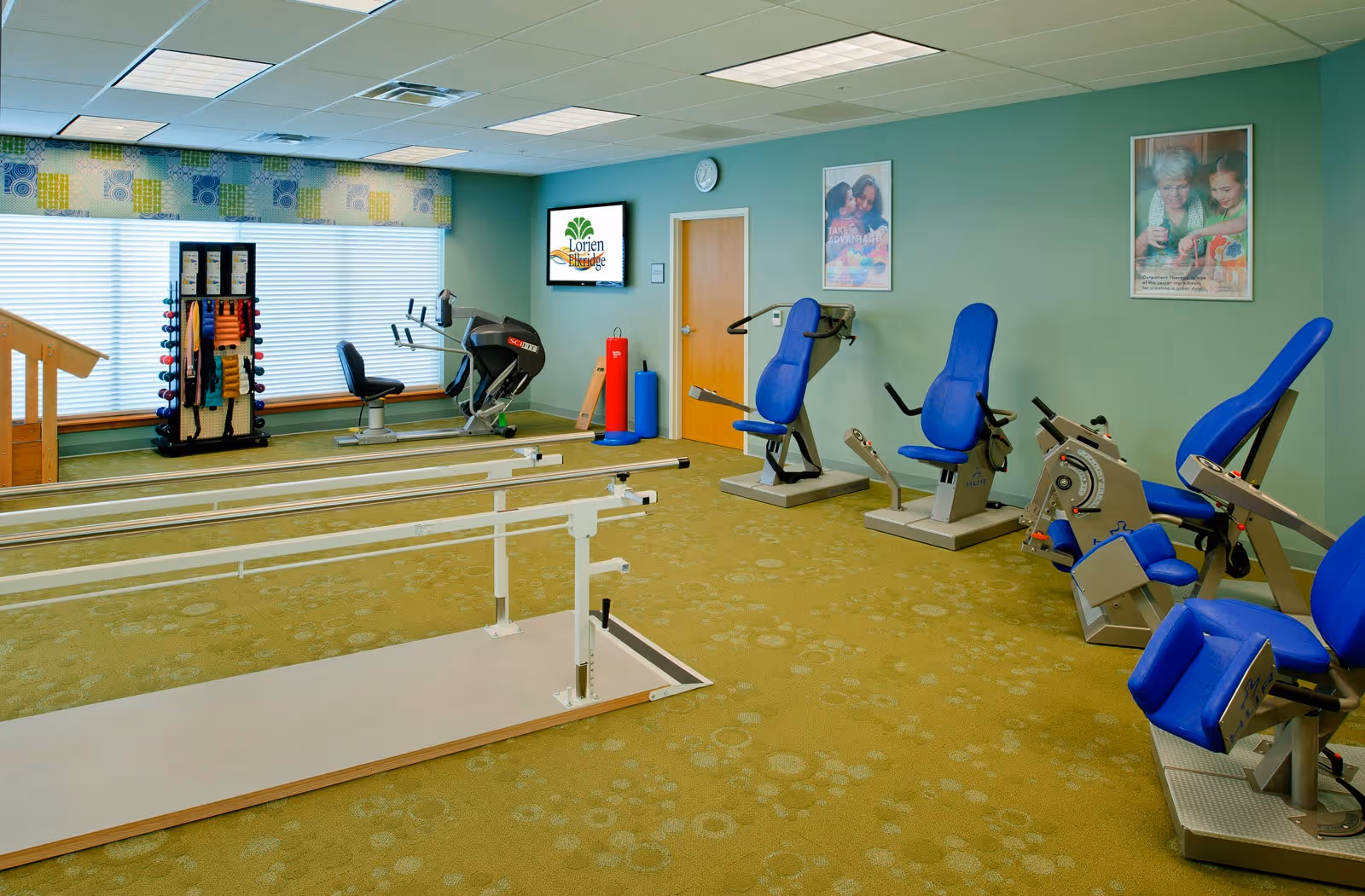 A senior living facility exercise room with various fitness equipment including blue padded exercise machines, a stationary bike, hand weights on a rack, and parallel bars for walking support. The room has green walls, a patterned green carpet, and large windows with patterned valances. There are two framed posters on the wall and a digital screen displaying the Lorien Elkridge logo.
