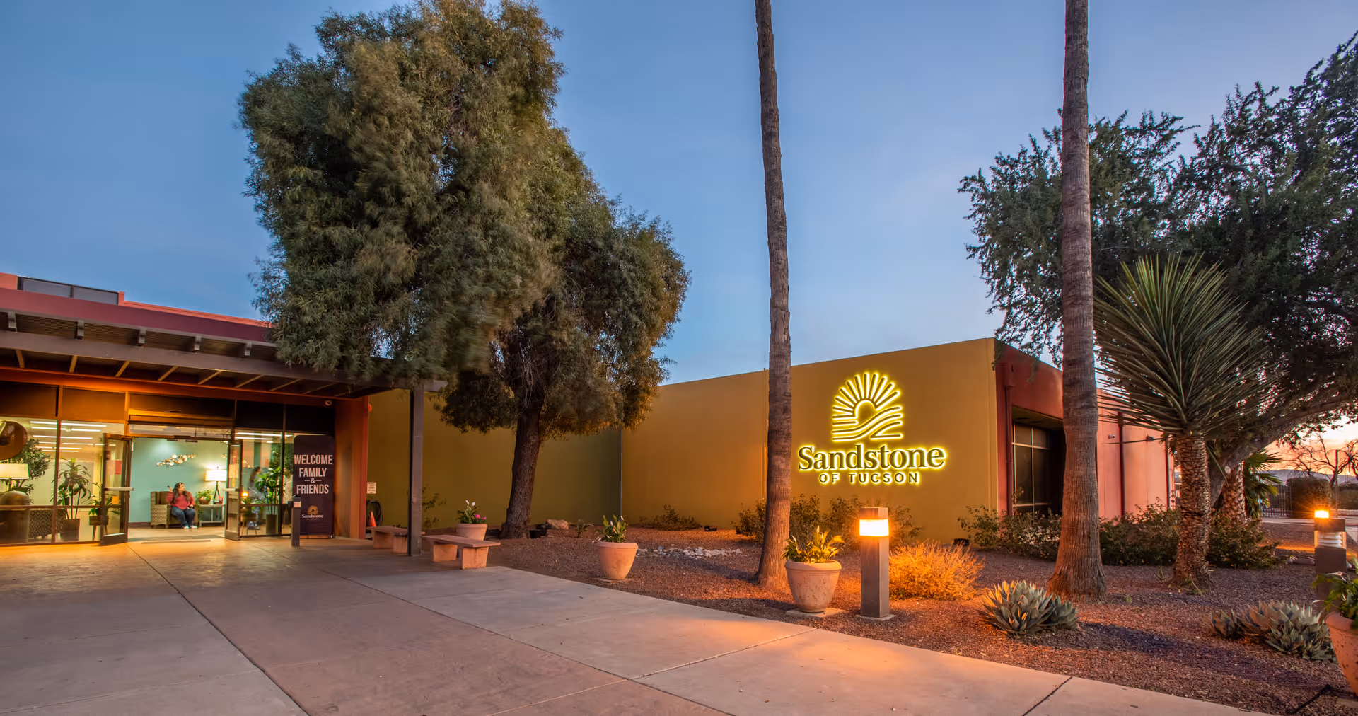 Front entrance of Sandstone of Tucson with an illuminated sign, landscaping, and a covered walkway at dusk.