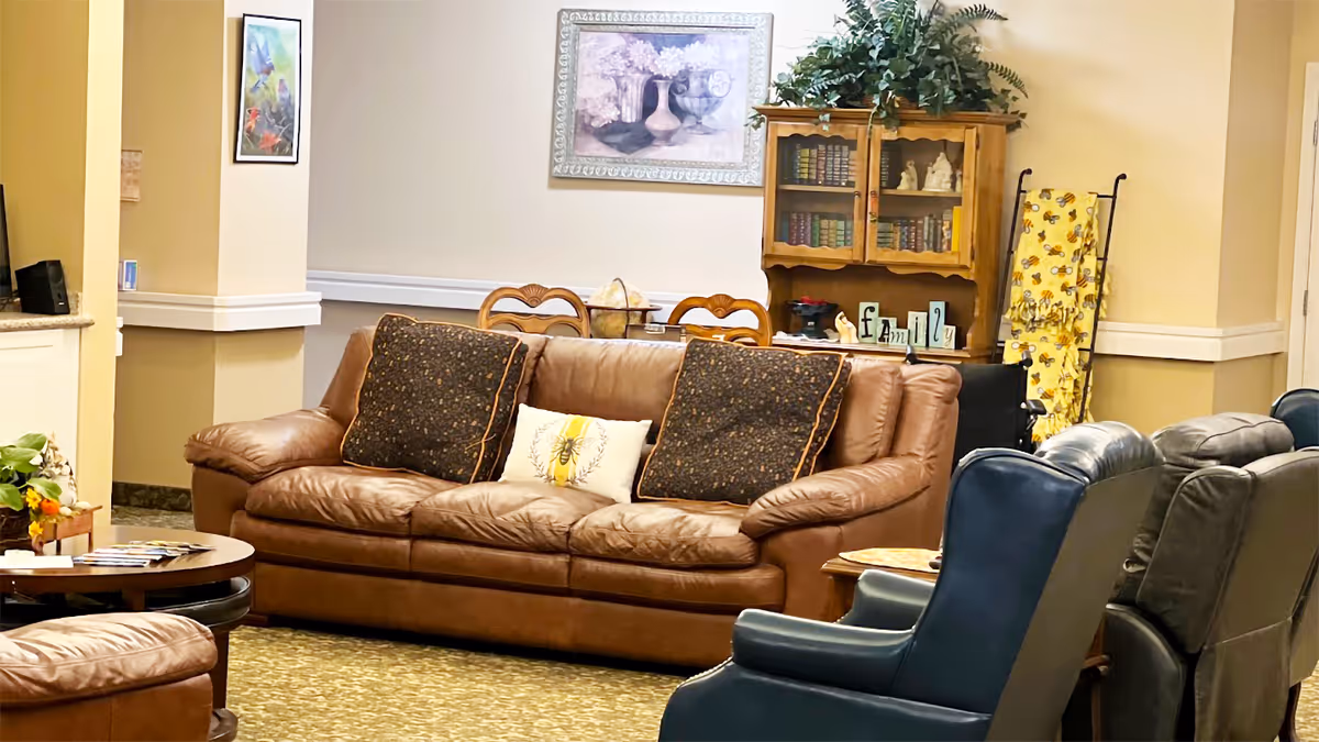 A cozy living room area with a brown leather sofa adorned with three decorative pillows, two dark patterned and one with a bee design. In front of the sofa are several blue and gray armchairs. Behind the sofa is a wooden dining table with chairs, a wooden cabinet filled with books and decorative items, and a framed floral painting on the wall. The room has warm beige walls and a patterned carpet floor.