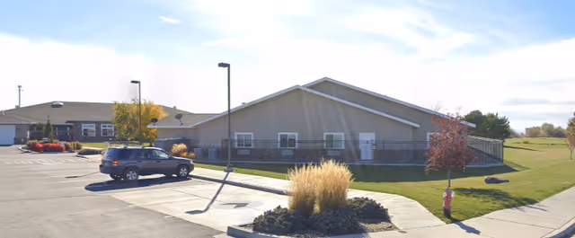 Exterior view of a single-story assisted living facility building with beige siding and a sloped roof. There is a parking lot with a parked SUV, a street lamp, landscaped areas with grass, bushes, and a small tree, and a fire hydrant near the sidewalk.