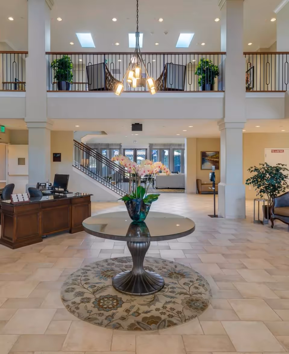 Spacious two-story lobby with a central round table holding an orchid arrangement, a reception desk, staircase, and seating on the upper level.