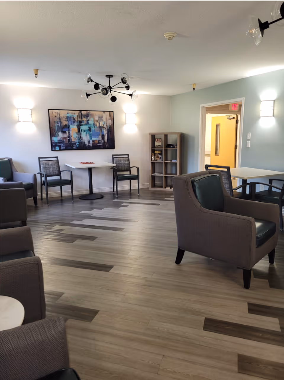 A well-lit common area with modern furniture including armchairs and tables. The room has a light-colored wooden floor with a pattern, a bookshelf with some items, and a colorful abstract painting on the wall. There are wall-mounted lights and a contemporary ceiling light fixture. An open doorway leads to a hallway with a yellow door and an exit sign above it.