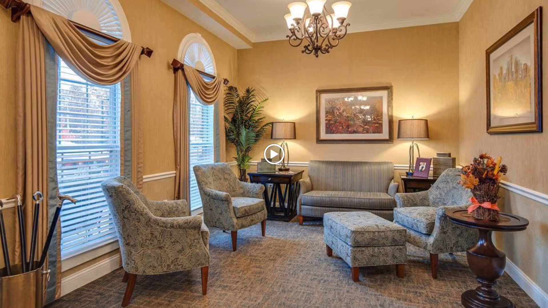 A cozy living room with three patterned armchairs, a matching ottoman, and a striped sofa. The room features beige walls, two large windows with blinds and brown drapes, two side tables with lamps, a round wooden table with a fall-themed floral arrangement, framed artwork on the walls, a chandelier, and a potted plant in the corner.