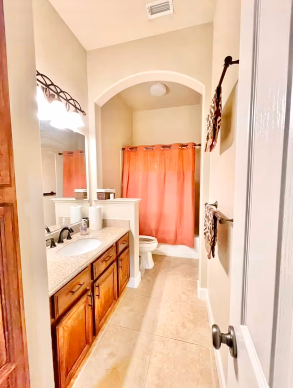 A bathroom with a wooden vanity featuring a granite countertop and a sink. Above the vanity is a large mirror with a light fixture. The bathroom has beige walls and tiled floor. At the far end, there is a toilet next to a bathtub with a coral-colored shower curtain. Two patterned towels hang on the wall to the right.