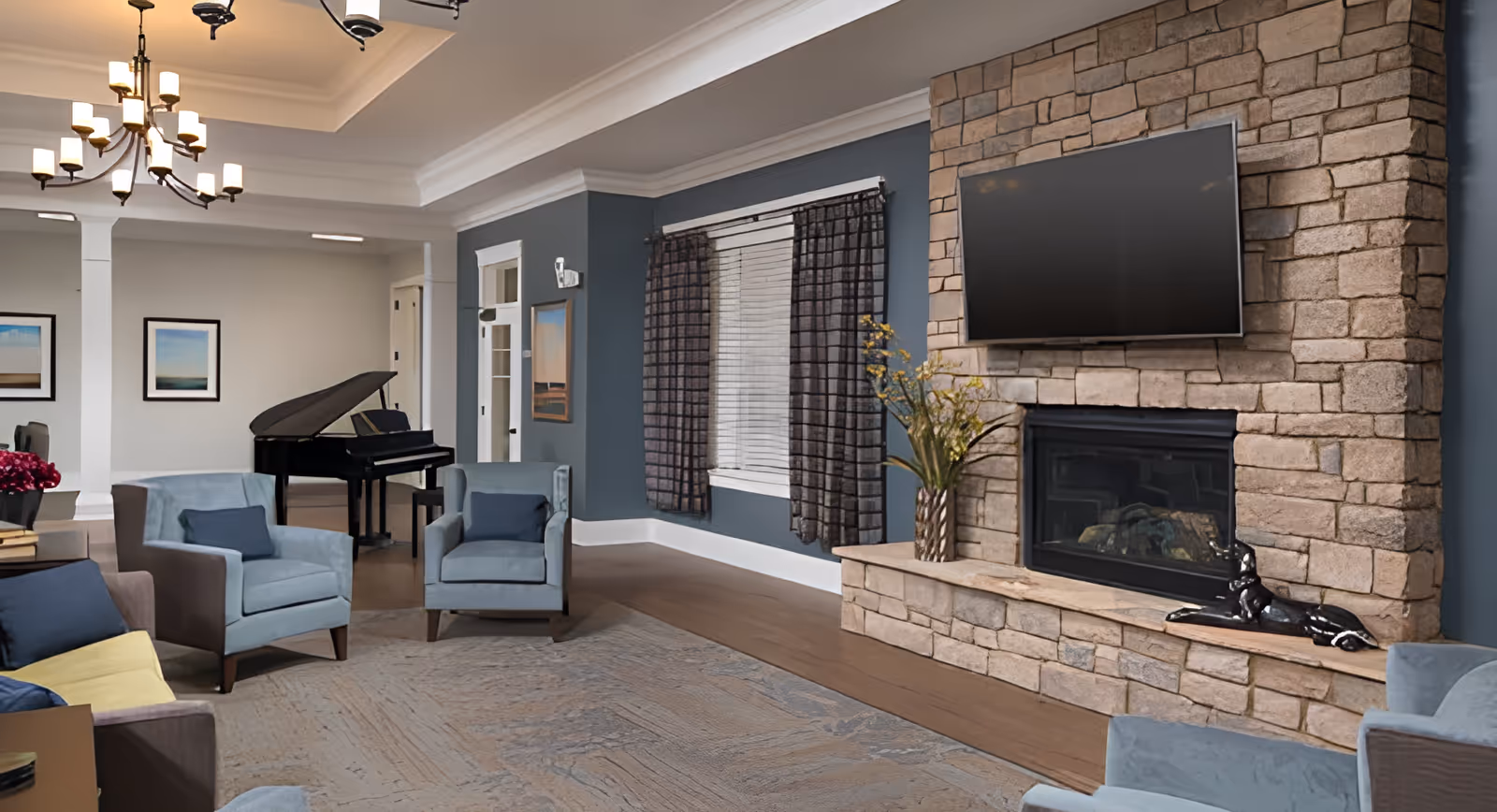 A cozy senior living facility common area with blue armchairs and a sofa arranged around a large stone fireplace with a mounted flat-screen TV. The room features a chandelier, framed artwork on the walls, a window with dark curtains, and a black grand piano in the background.
