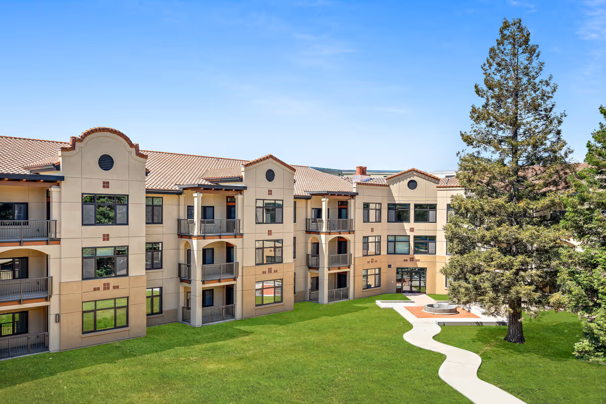 Exterior view of The Meadows of Napa Valley senior living facility showing a three-story building with balconies, large windows, a tiled roof, and a well-maintained green lawn with a curved concrete pathway and a large tree near a circular seating area.