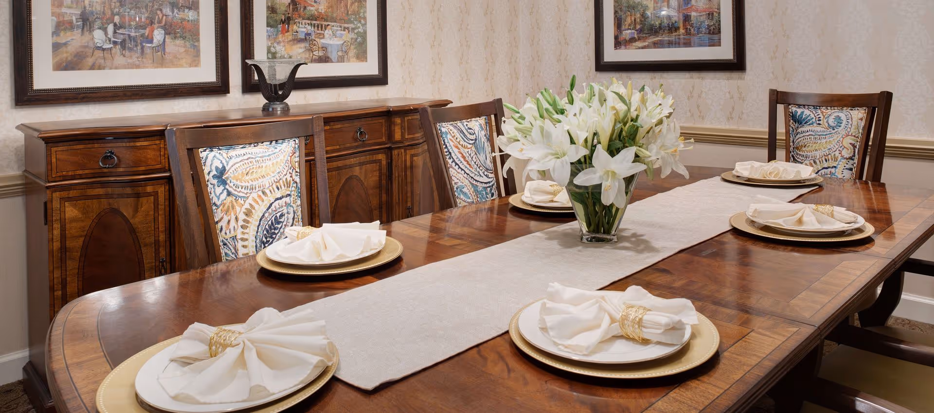 A formal dining room with a polished wooden table set for five people. Each place setting includes a gold charger plate, a white plate, and a neatly folded white napkin with a gold napkin ring. A beige table runner runs down the center of the table, topped with a clear vase filled with white lilies. The chairs have wooden frames with patterned fabric backs. In the background, there is a wooden sideboard with decorative items and framed artwork on the wall.