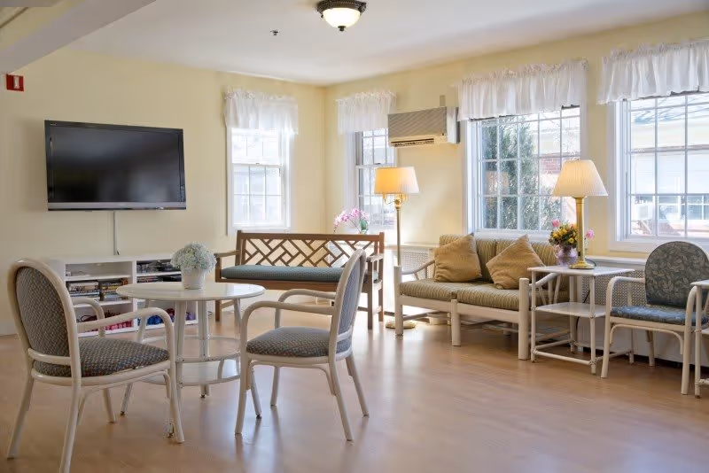 A bright and cozy living room area in a senior living facility with light-colored walls and wooden flooring. The room features a wall-mounted flat-screen TV, a small round table with four cushioned chairs, a wooden bench with a cushion, a cushioned loveseat with pillows, two side tables with lamps, and several windows letting in natural light. There are also flower arrangements on the tables.