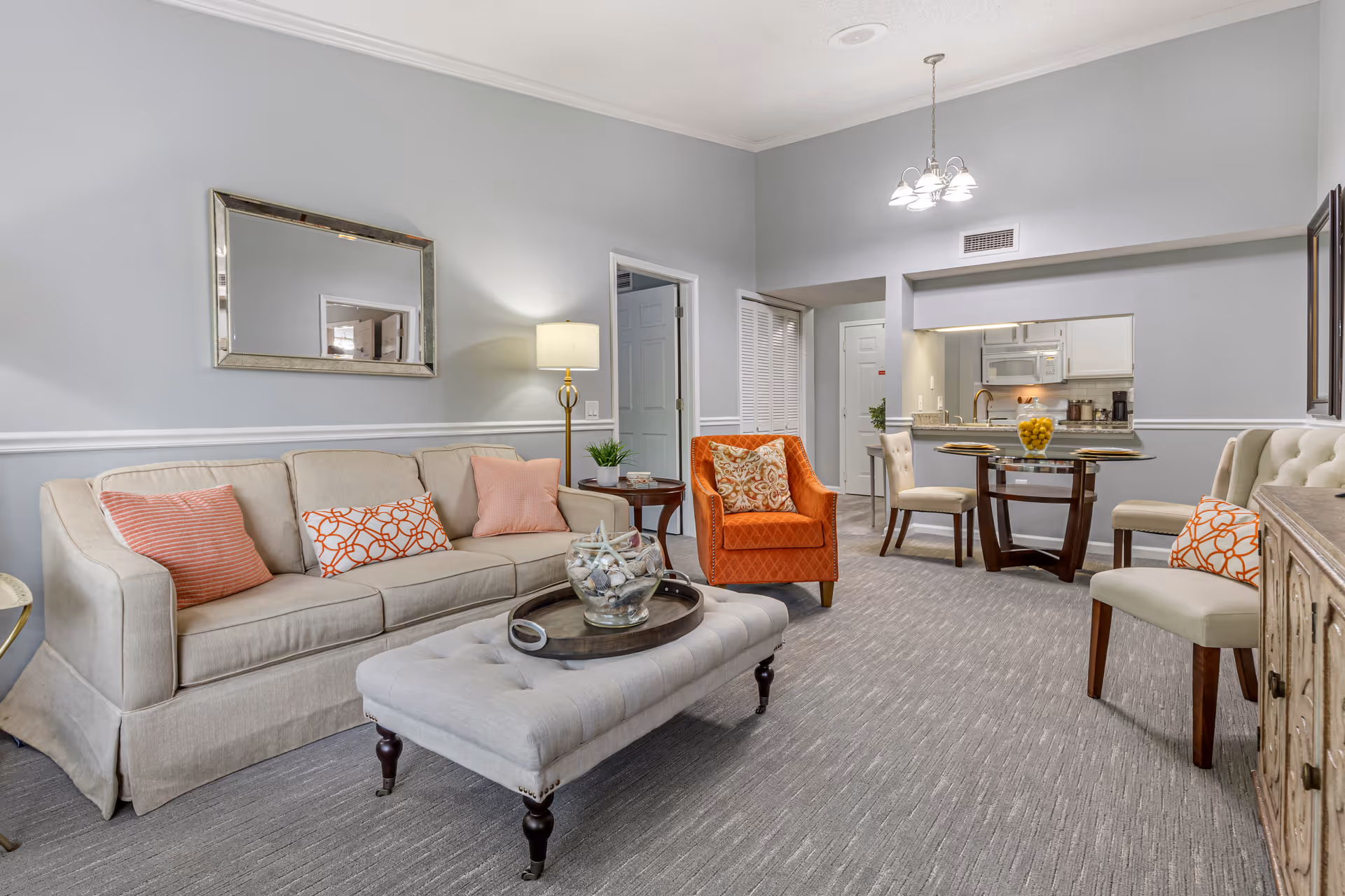 Well-lit living room with a beige sofa, tufted ottoman, orange armchair, and a small dining table visible in the background.