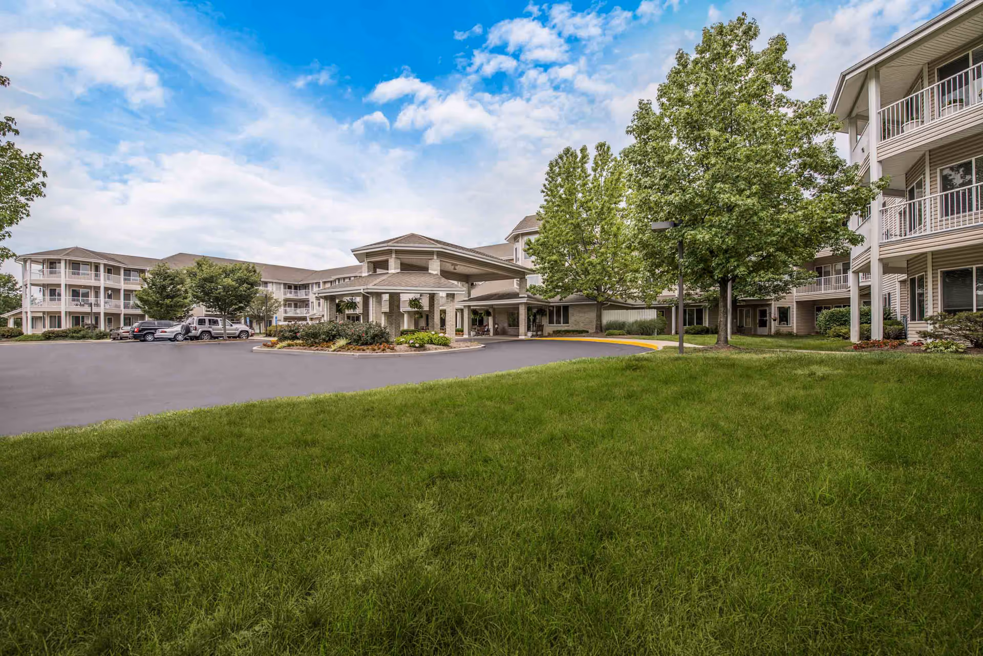 Exterior front entrance of a senior living building with a circular driveway, parked cars, green lawn and trees under a blue sky.