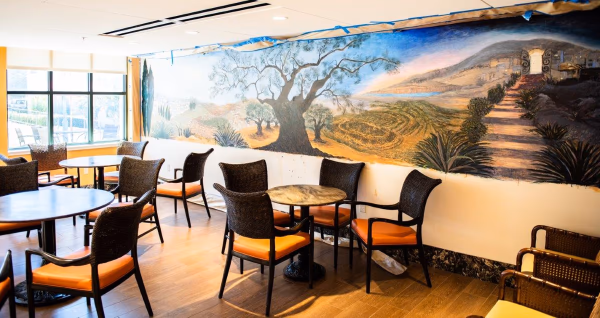 A bright dining area with several round tables and wicker chairs with orange cushions. The room features a large mural on the wall depicting a scenic landscape with trees, hills, and a pathway leading to a gate. Large windows allow natural light to fill the space.