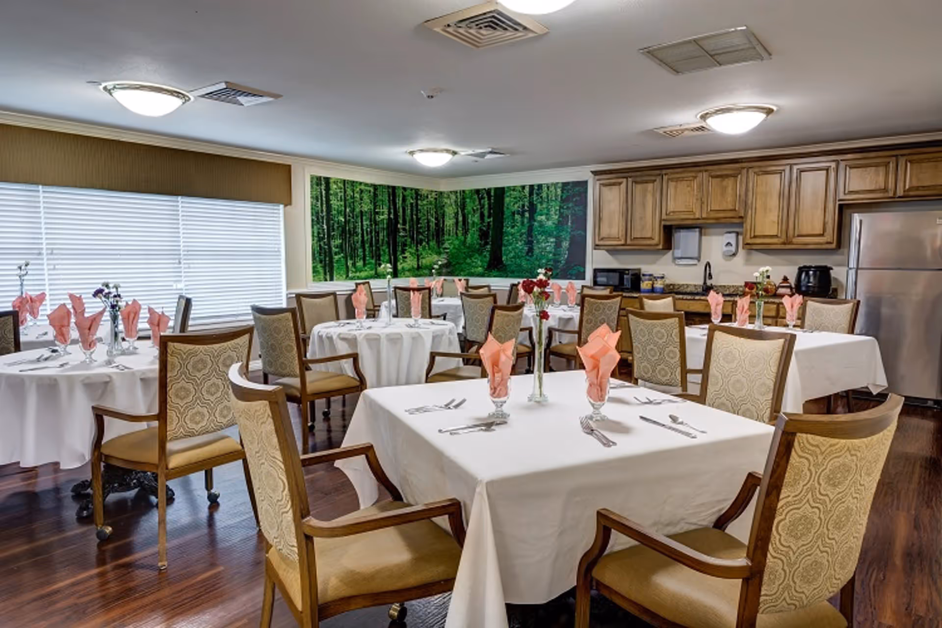A dining room with several tables covered in white tablecloths, each set with folded pink napkins, silverware, and small flower vases. The room has wooden chairs with patterned upholstery, wood flooring, and a kitchen area with wooden cabinets and stainless steel appliances in the background. A large wall mural of a green forest adds a natural touch to the space.