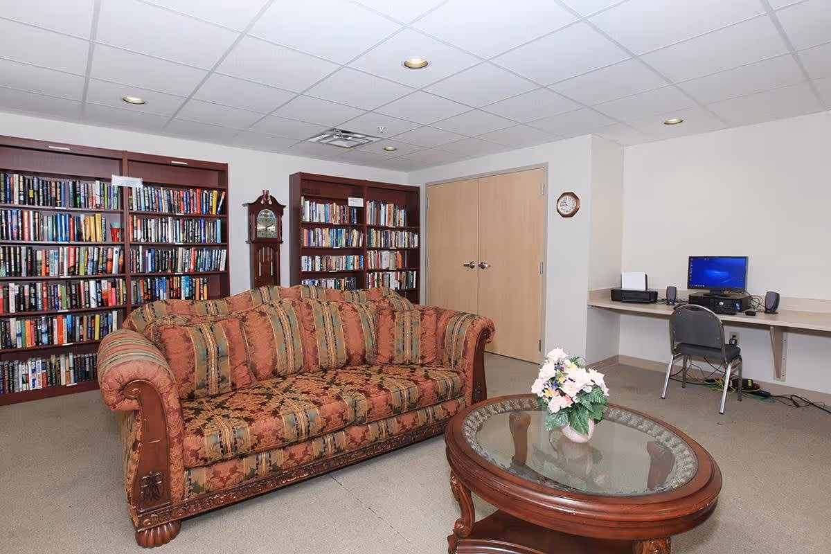 A cozy room with a patterned sofa in the center, a round glass-top wooden coffee table with a flower arrangement, two large bookshelves filled with books, a grandfather clock between the bookshelves, a wall clock, and a computer workstation with a desktop computer, printer, and chair against the wall.