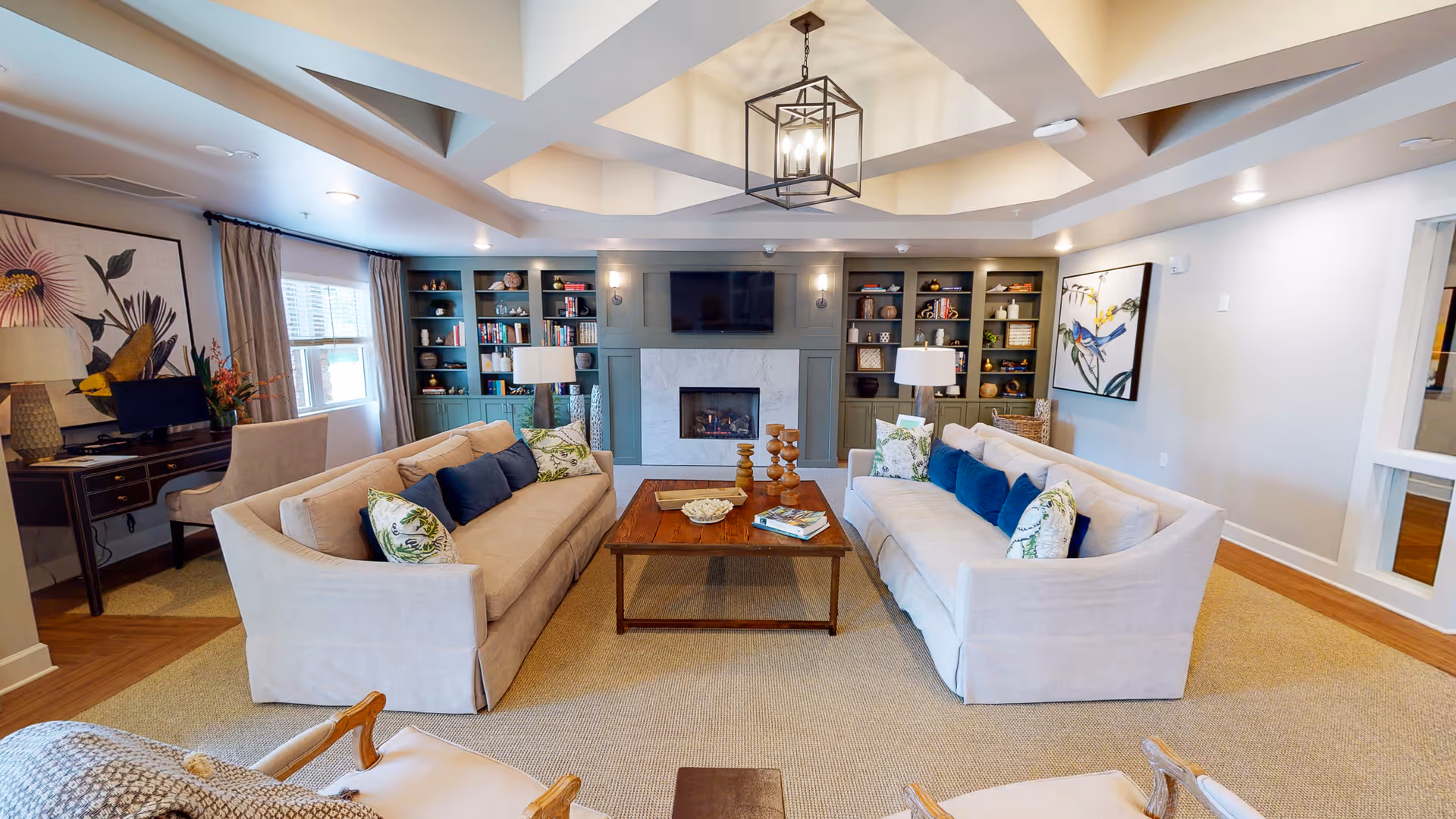 A spacious and well-lit living room with two beige sofas facing each other across a wooden coffee table. The room features a modern fireplace with built-in shelves on either side filled with decorative items and books. There are two table lamps on side tables next to the sofas, large windows with curtains, and framed artwork on the walls. The ceiling has a recessed design with a central hanging light fixture.