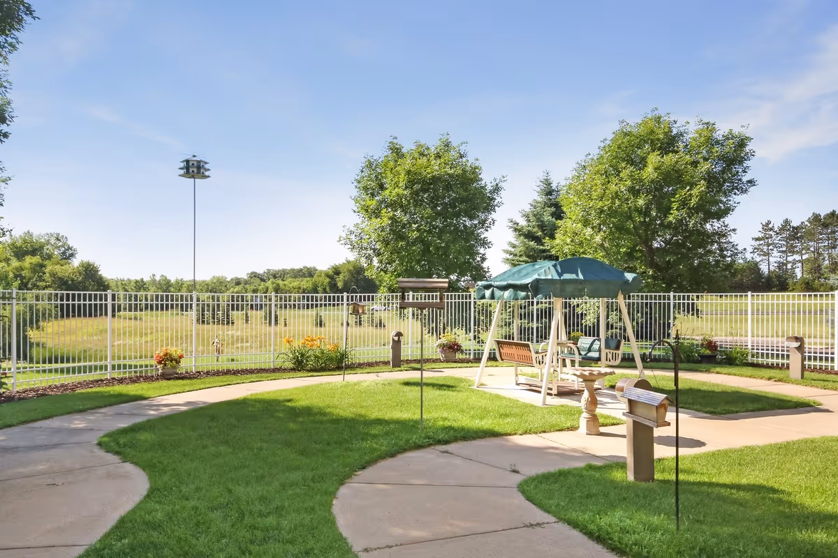 Outdoor garden area with a curved concrete pathway, green grass, several bird feeders, a white metal fence, and a covered swing bench under a green canopy. Trees and open fields are visible in the background under a clear blue sky.