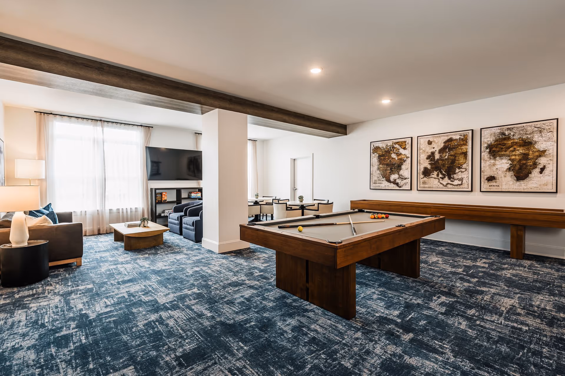 A spacious senior living common area with a pool table in the foreground, a shuffleboard table against the wall, a seating area with a sofa, armchairs, coffee table, and a large TV mounted on the wall. The room has large windows with sheer curtains, blue patterned carpet, and three framed vintage world maps on the wall.