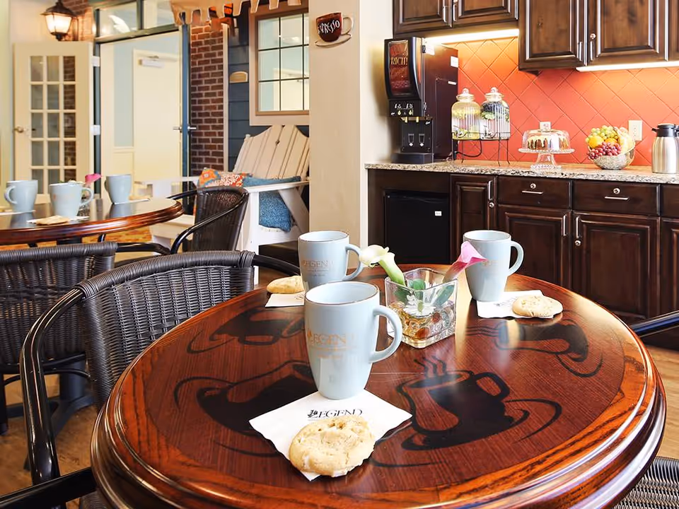 A cozy dining area with round wooden tables featuring coffee cup designs, each table set with light blue mugs and cookies on napkins. In the background, there is a kitchen counter with dark wooden cabinets, a coffee machine, glass jars, a fruit bowl, and a cake stand. The setting has a warm and inviting atmosphere.