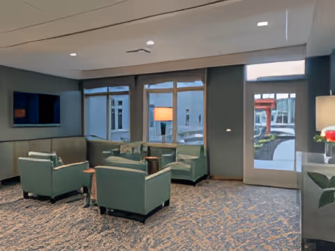 A cozy living room area with green upholstered armchairs and a matching sofa arranged around a small wooden coffee table. Large windows and a glass door let in natural light, and a floor lamp is visible near the seating. The carpet has a patterned design, and the walls are painted in a muted green color.